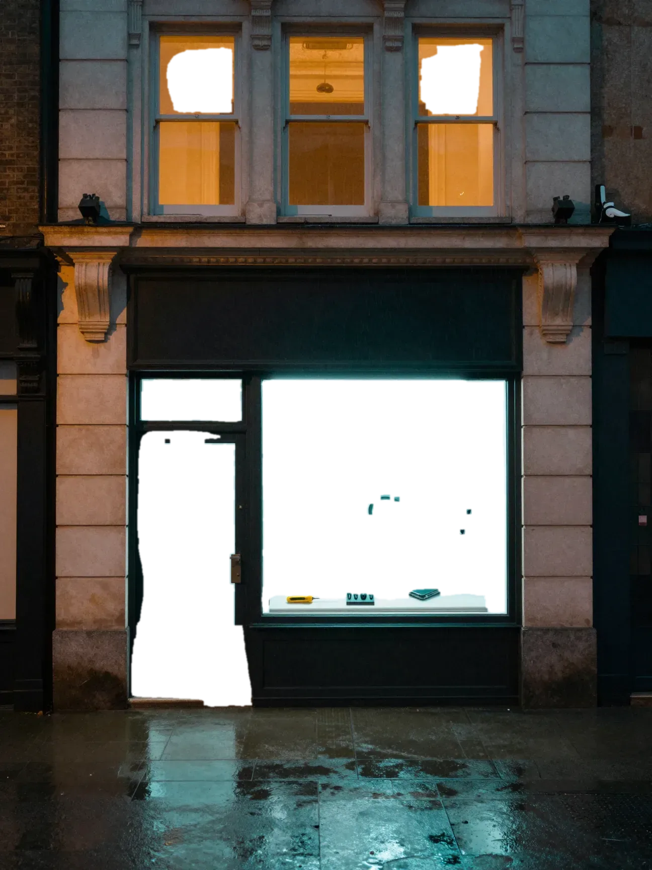 A pale limestone storefront at night. Under a white light, the shop window shows two smartphones on clear stands, a magnifier lamp and a row of precision screwdrivers.